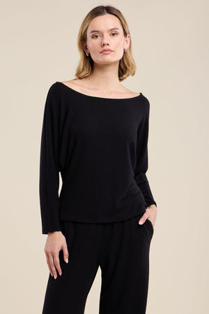 A person wearing the CECILIA TOP by Velvet by Graham & Spencer— a black, relaxed fit long-sleeve top —and black pants stands against a plain beige background, facing forward with a neutral expression.