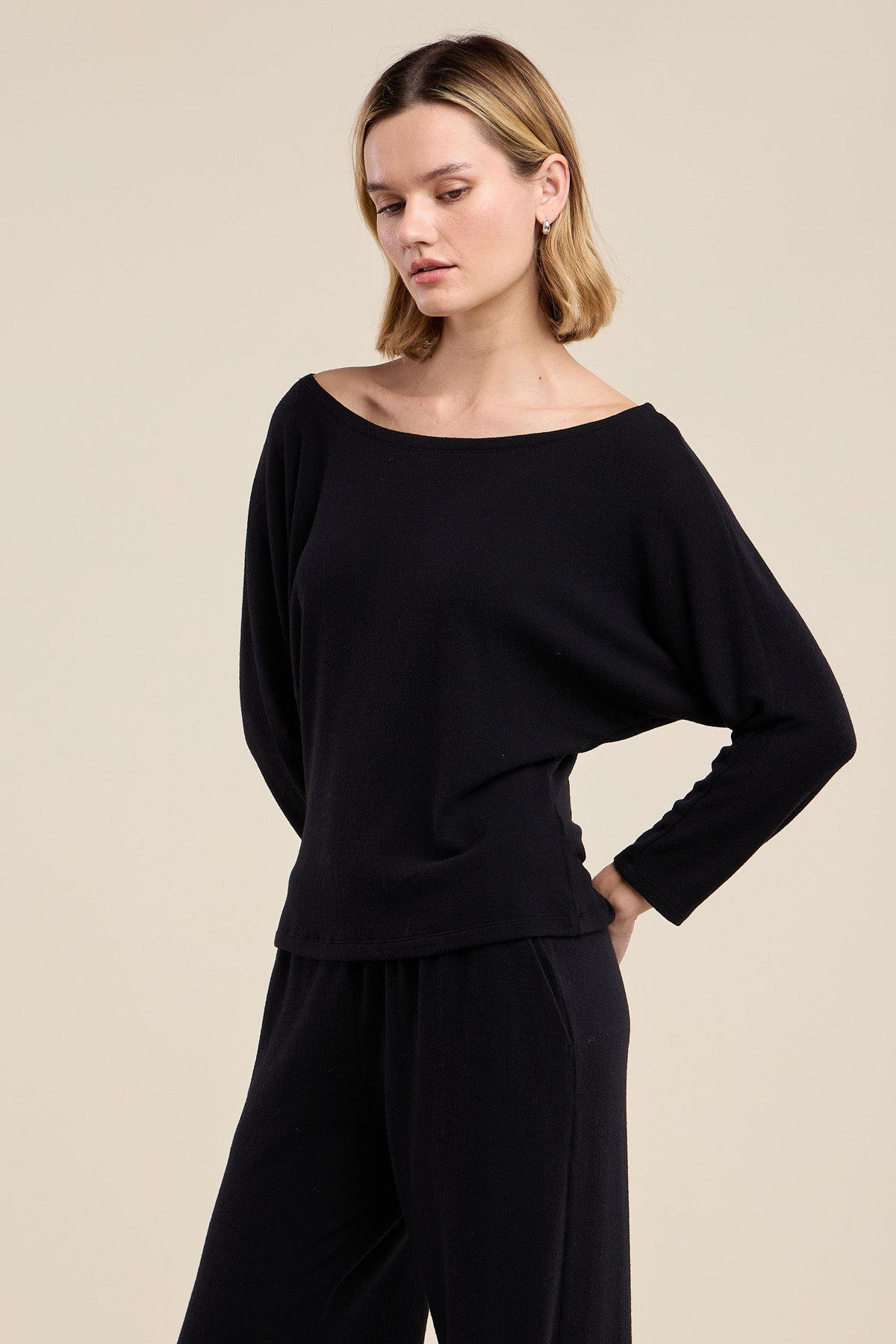 Wearing the CECILIA TOP by Velvet by Graham & Spencer, a woman stands against a plain beige background in a relaxed pose, dressed in a matching black ensemble with one hand on her hip and her gaze turned downward.