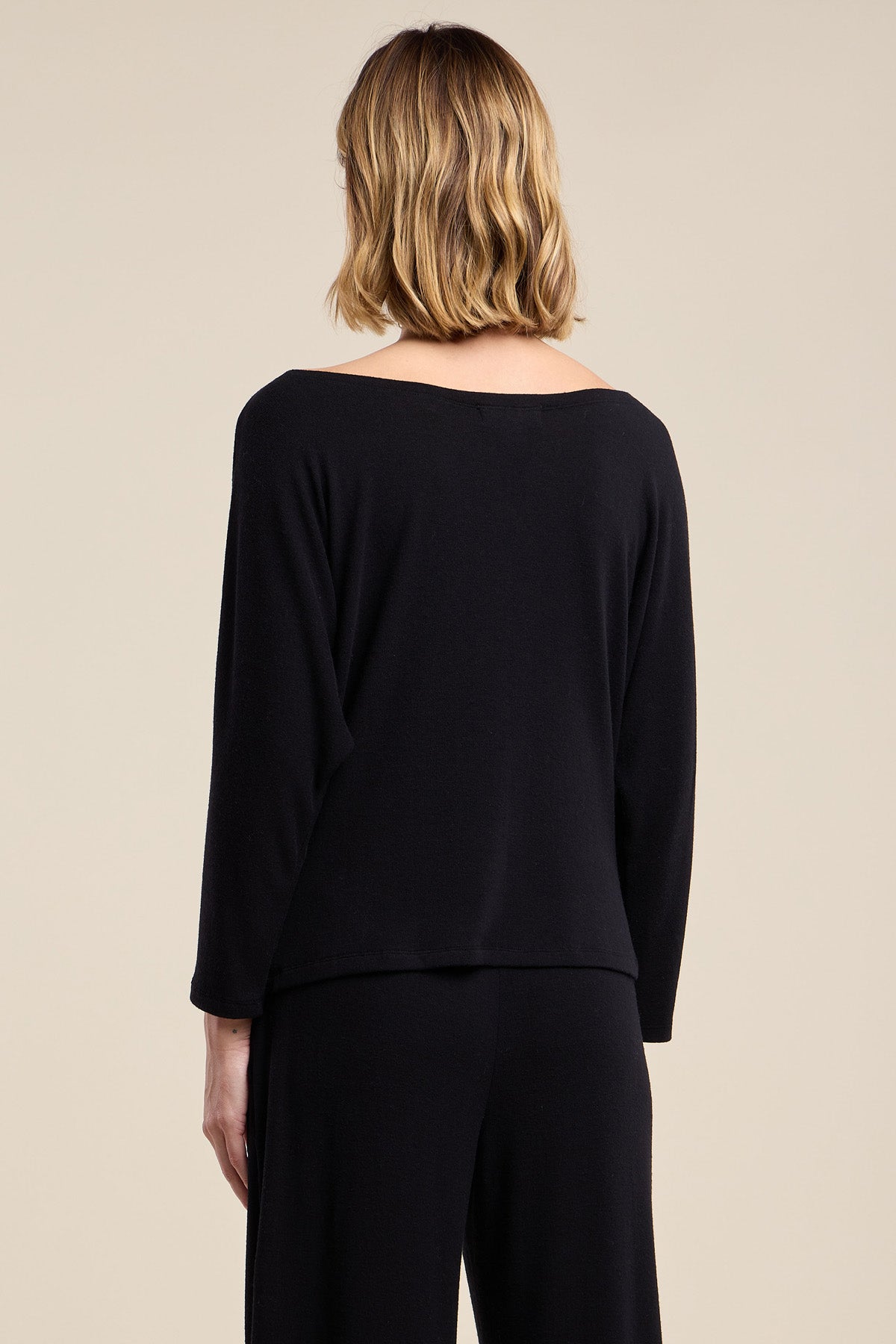 A person with shoulder-length blonde hair is seen from the back against a plain beige background, wearing the Velvet by Graham & Spencer CECILIA TOP—a relaxed fit, soft knit black long-sleeve top—paired with black pants.-42063736864961