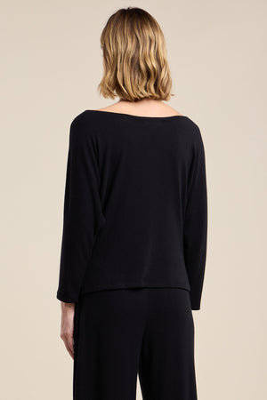 A person with shoulder-length blonde hair is seen from the back against a plain beige background, wearing the Velvet by Graham & Spencer CECILIA TOP—a relaxed fit, soft knit black long-sleeve top—paired with black pants.
