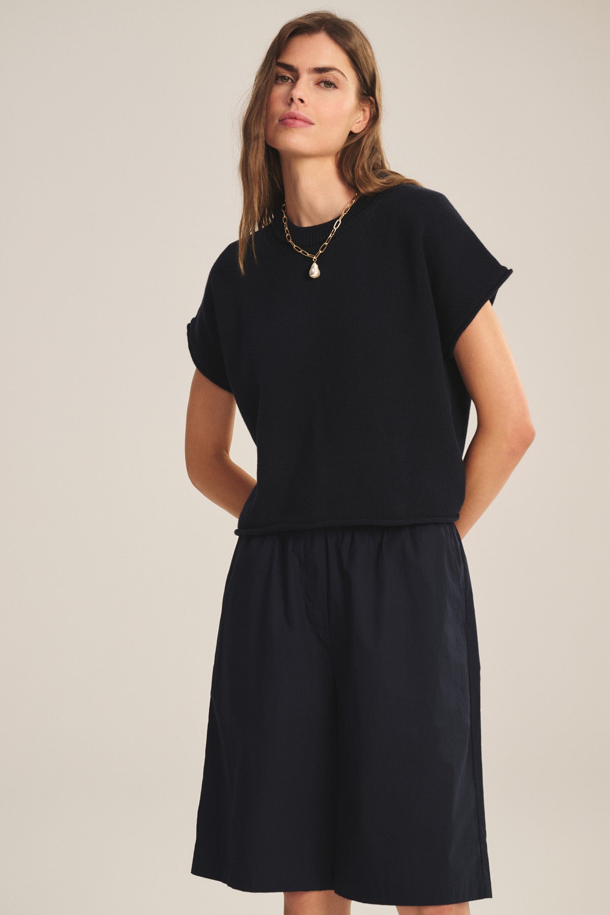 A woman stands against a plain background wearing the KEENY SWEATER by Velvet by Graham & Spencer—a black cotton-cashmere short-sleeve top with a boxy silhouette—paired with black knee-length shorts and a gold necklace.