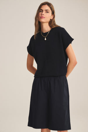 A woman stands against a plain background wearing the KEENY SWEATER by Velvet by Graham & Spencer—a black cotton-cashmere short-sleeve top with a boxy silhouette—paired with black knee-length shorts and a gold necklace.
