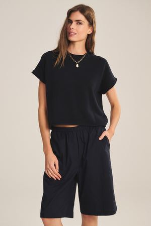 A woman wears the KEENY SWEATER by Velvet by Graham & Spencer—a black cotton-cashmere, boxy short-sleeve top—paired with black knee-length shorts and a necklace, standing with one hand in her pocket against a plain background.