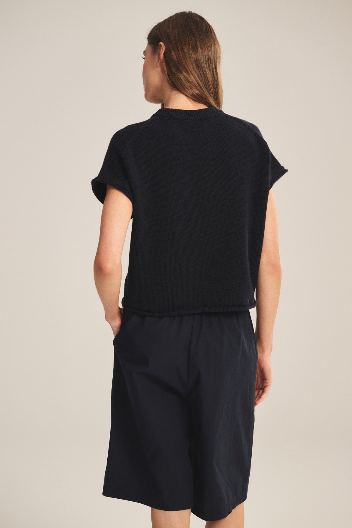 A person with long brown hair stands facing away, wearing the KEENY SWEATER by Velvet by Graham & Spencer and knee-length black shorts against a neutral background.