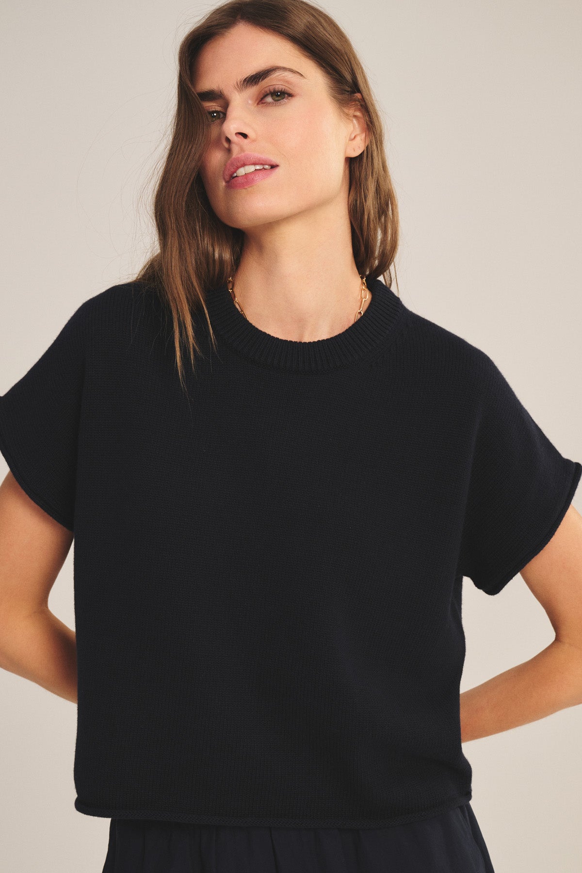 A woman with long brown hair wears the KEENY SWEATER by Velvet by Graham & Spencer—a short-sleeved, black cotton-cashmere knit top with a boxy silhouette and gold necklace—standing against a plain beige background.-41038337736897