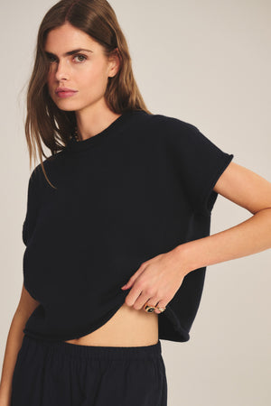 A woman with long brown hair wears the KEENY SWEATER by Velvet by Graham & Spencer—a cropped, boxy cotton-cashmere top—paired with black pants, standing against a neutral background and lifting the hem slightly.