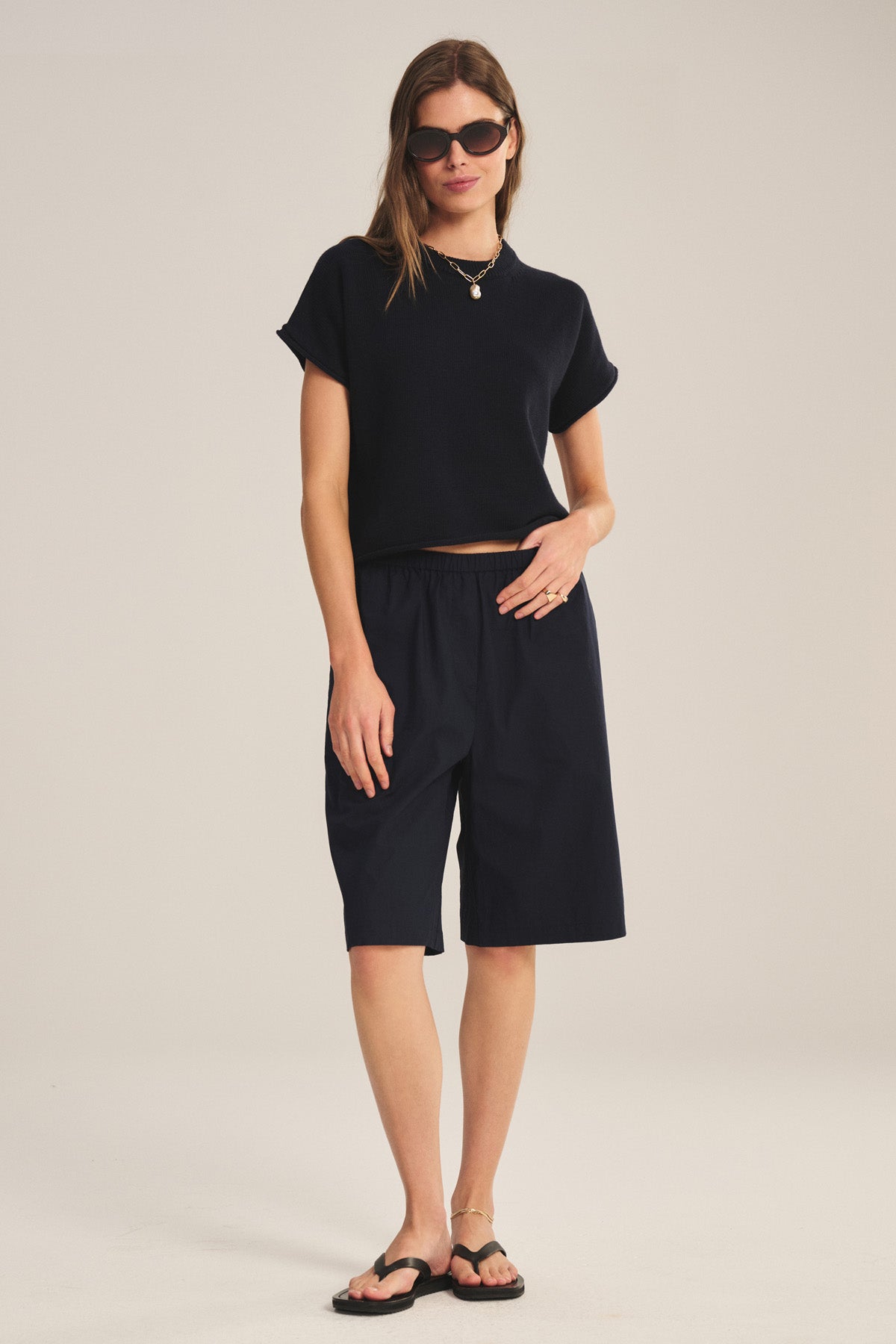 A woman stands in a studio wearing the KEENY SWEATER by Velvet by Graham & Spencer, paired with sunglasses, black knee-length shorts, and black sandals.