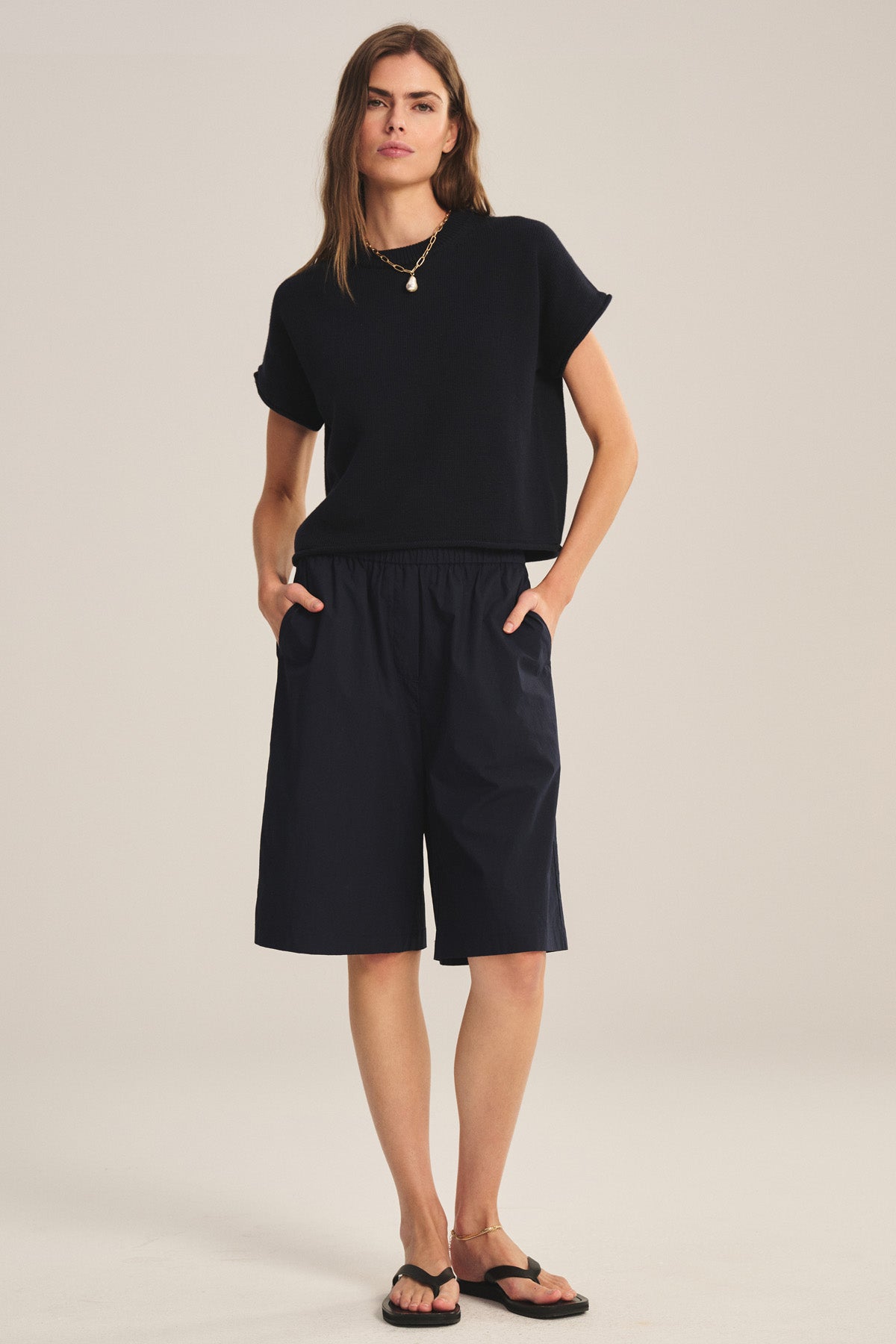 A woman stands against a plain background, hands in pockets, wearing the KEENY SWEATER by Velvet by Graham & Spencer—a black short-sleeve boxy top—paired with loose black knee-length shorts, black sandals, and a necklace.-41038337638593