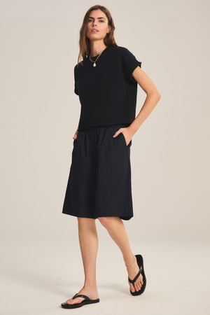 A woman stands against a plain background wearing the KEENY SWEATER by Velvet by Graham & Spencer—a black cotton-cashmere, boxy sweater—paired with black knee-length shorts, black sandals, layered necklaces, and her hands in her pockets.