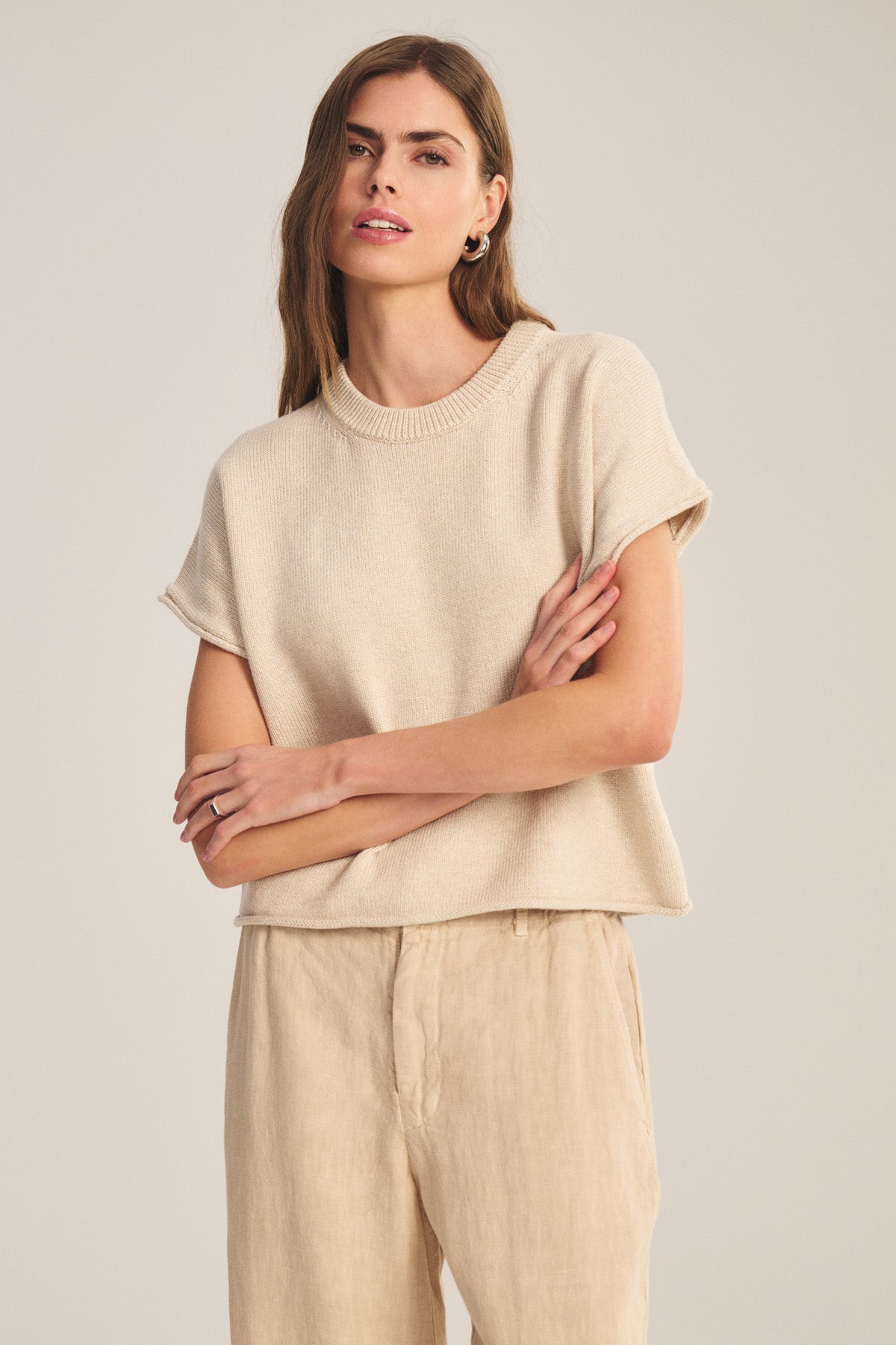 A woman stands with arms crossed, wearing the KEENY SWEATER by Velvet by Graham & Spencer and light beige pants against a plain, neutral background.