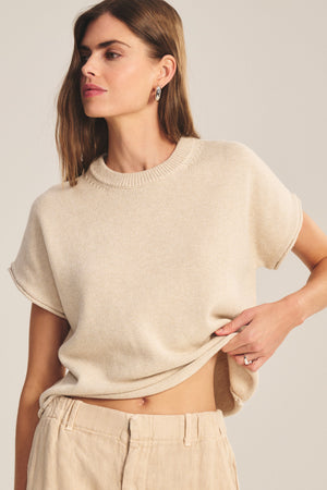 A woman wears the KEENY SWEATER by Velvet by Graham & Spencer—a short-sleeve, beige, boxy cropped sweater with matching pants—holding up the hem with one hand.