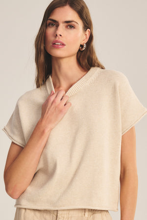 A woman with long brown hair wears the KEENY SWEATER by Velvet by Graham & Spencer, a soft beige knit cropped sweater paired with light-colored pants, standing against a neutral background.