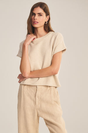 A woman poses against a neutral background in the KEENY SWEATER by Velvet by Graham & Spencer, a cropped, short-sleeve cotton-cashmere top, paired with light tan pants and one arm bent with her hand near her chin.