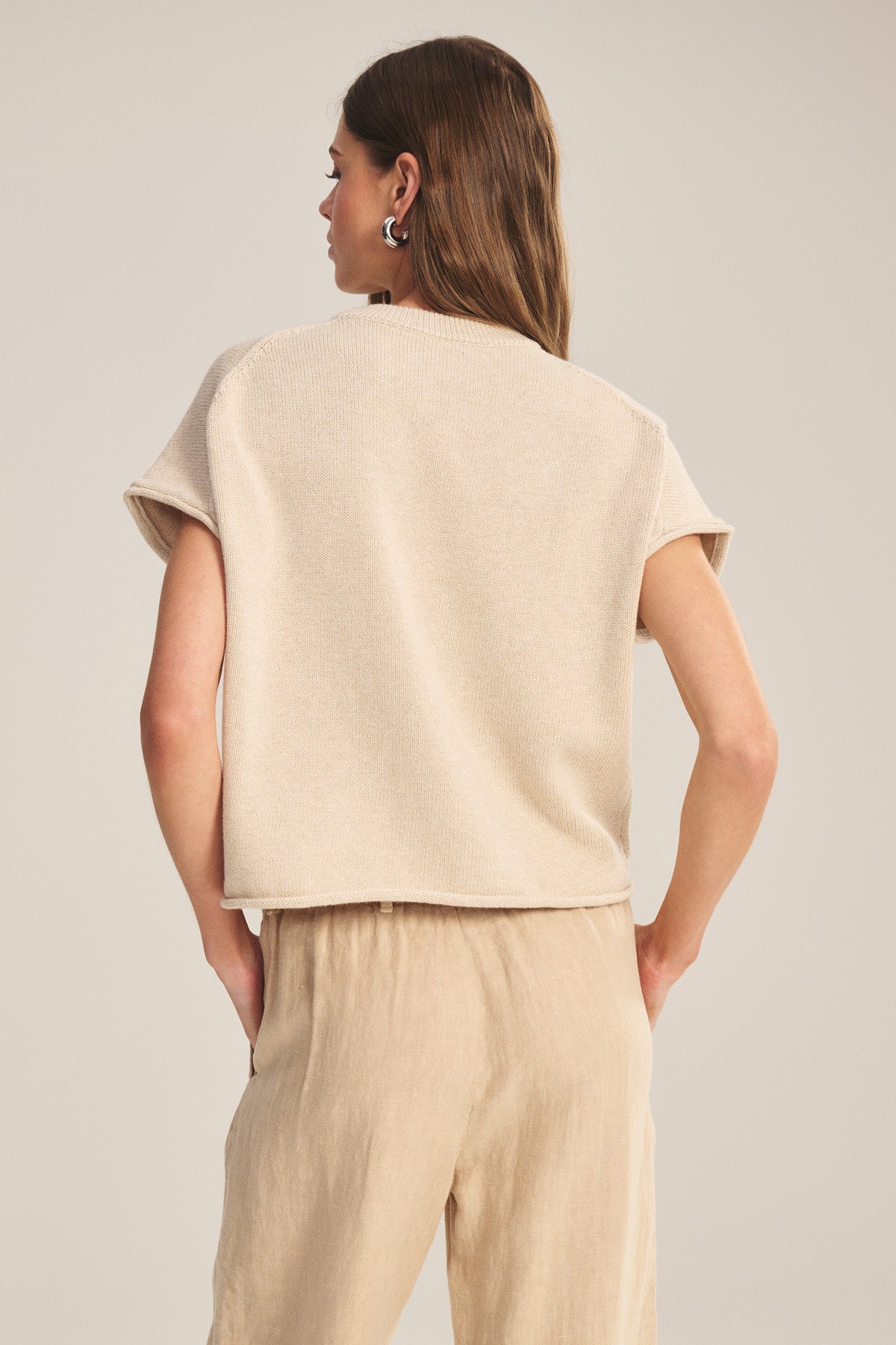 A woman faces away, wearing the KEENY SWEATER by Velvet by Graham & Spencer—a boxy short-sleeve beige top—paired with loose light tan pants against a plain background.