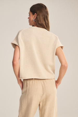 A woman faces away, wearing the KEENY SWEATER by Velvet by Graham & Spencer—a boxy short-sleeve beige top—paired with loose light tan pants against a plain background.