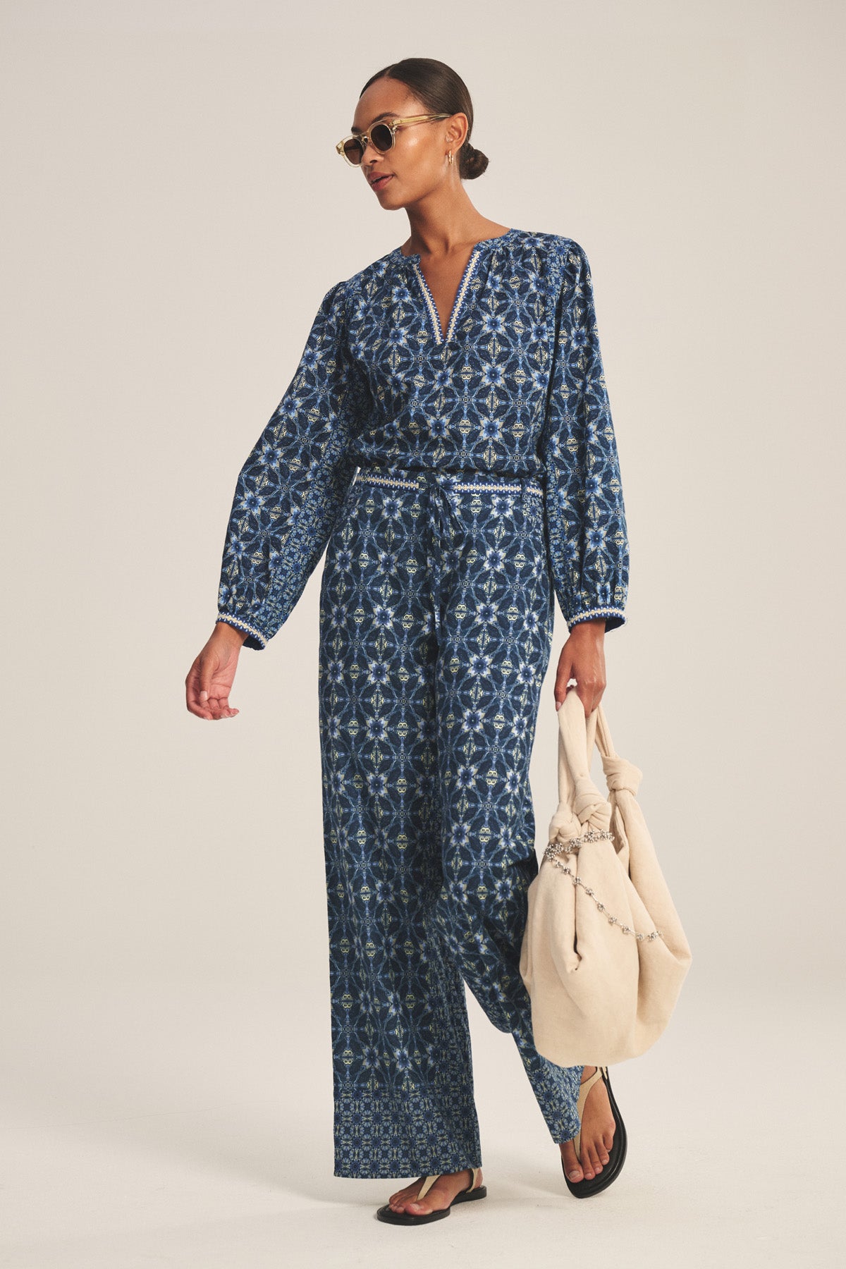A person wearing the KENDRA PANT by Velvet by Graham & Spencer, styled with black sandals, sunglasses, and a beige tote bag, walks against a plain background.-41008282009793