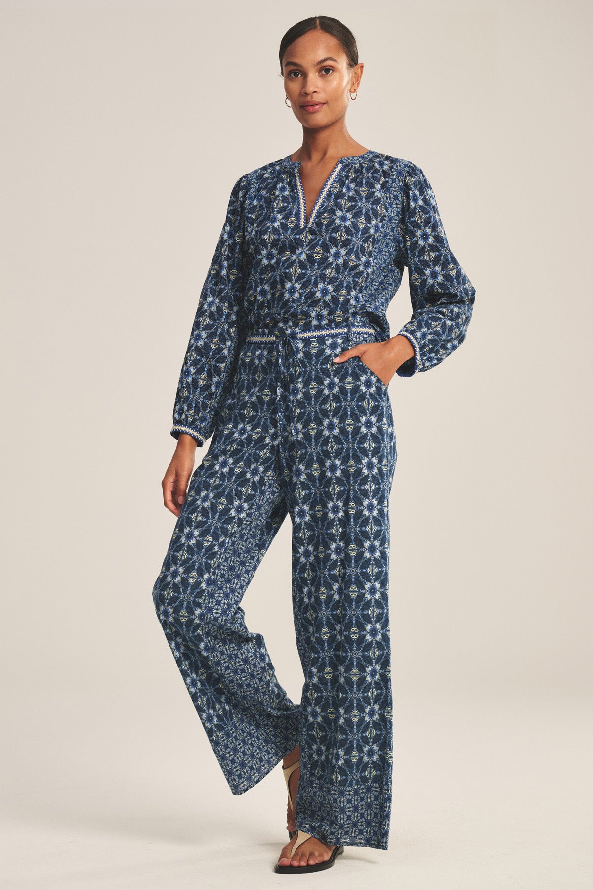 A woman wearing the Velvet by Graham & Spencer KENDRA PANT, a blue patterned wide-leg jumpsuit with long sleeves, a V-neckline, an embroidered belt, and one hand in her pocket.