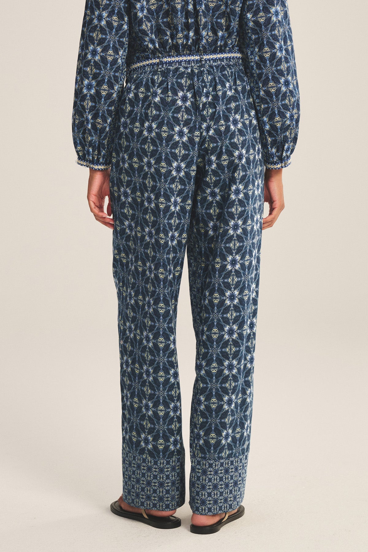 A person faces away, wearing the KENDRA PANT by Velvet by Graham & Spencer, a navy blue geometric-patterned jumpsuit with an embroidered belt and black sandals, set against a plain background.-41008281845953