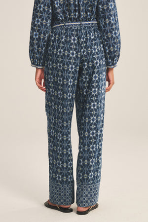 A person faces away, wearing the KENDRA PANT by Velvet by Graham & Spencer, a navy blue geometric-patterned jumpsuit with an embroidered belt and black sandals, set against a plain background.