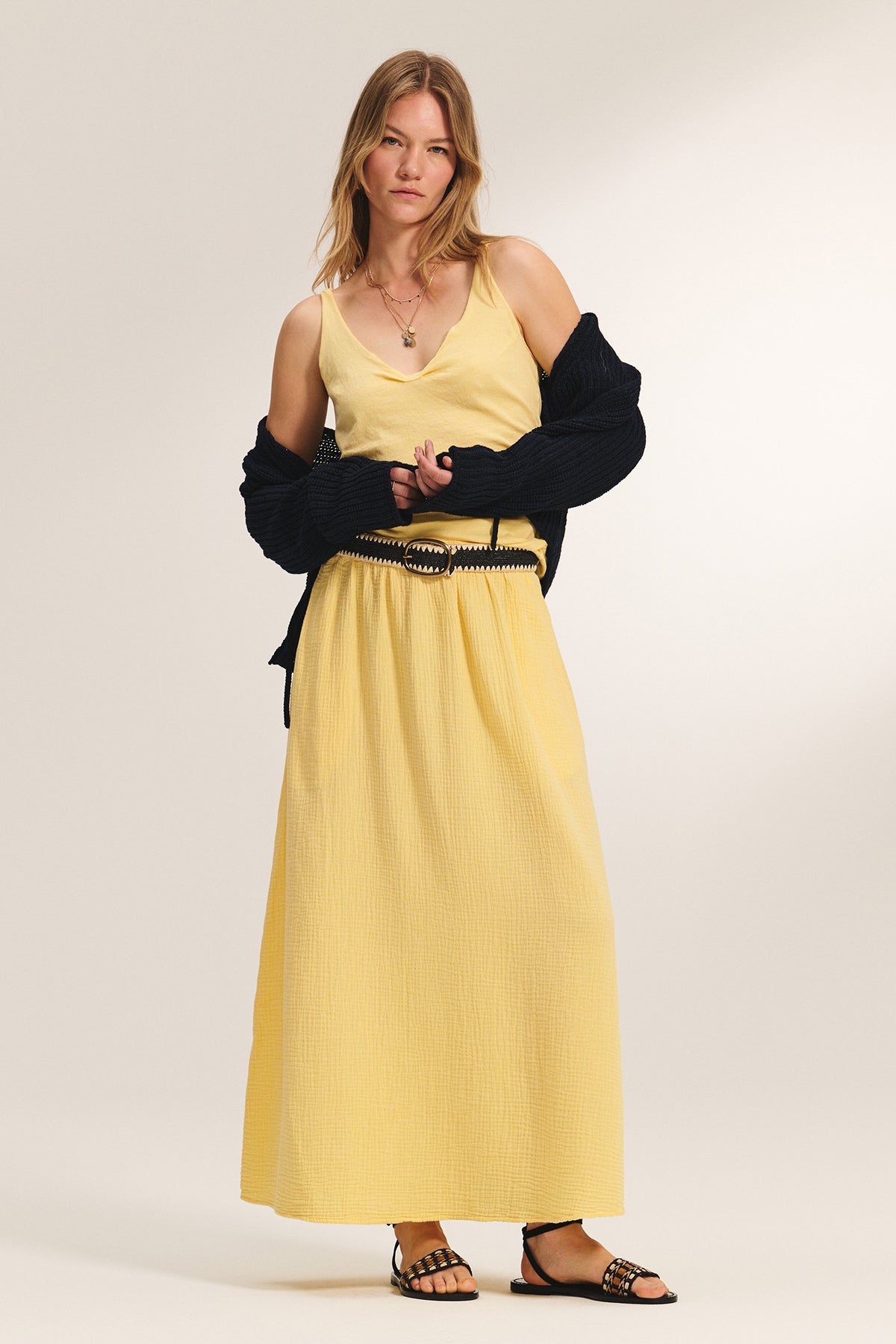 A woman stands against a plain background wearing the THALIA CARDIGAN by Velvet by Graham & Spencer draped over her arms, paired with a yellow sleeveless dress, black belt, and black sandals.-39292657369281