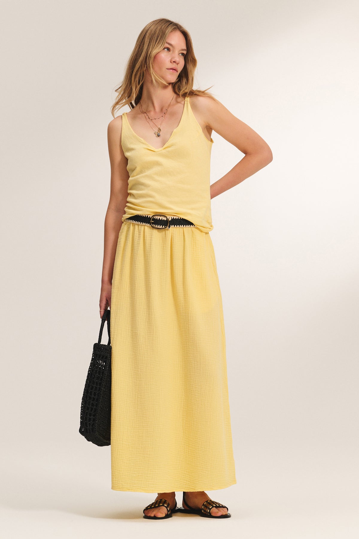 A woman stands against a plain background wearing the KIM TANK TOP by Velvet by Graham & Spencer—a lightweight cotton tank with a scoop neckline—paired with a matching long skirt, black belt, sandals, and a black woven bag for versatile style.-39173080252609