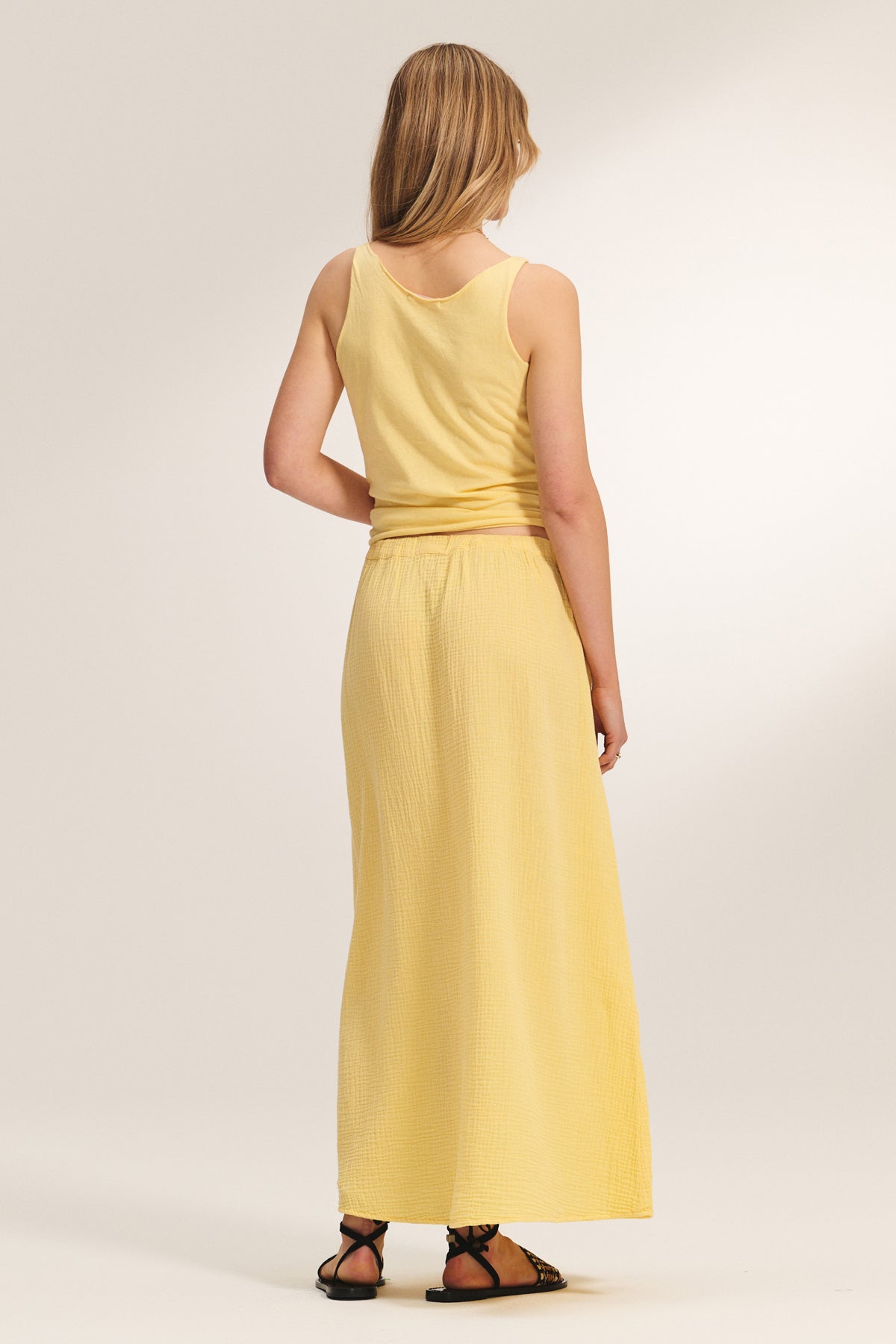 A woman faces away, wearing the sleeveless KIM TANK TOP by Velvet by Graham & Spencer and a long yellow skirt with black sandals, set against a plain light background.-39173077795009