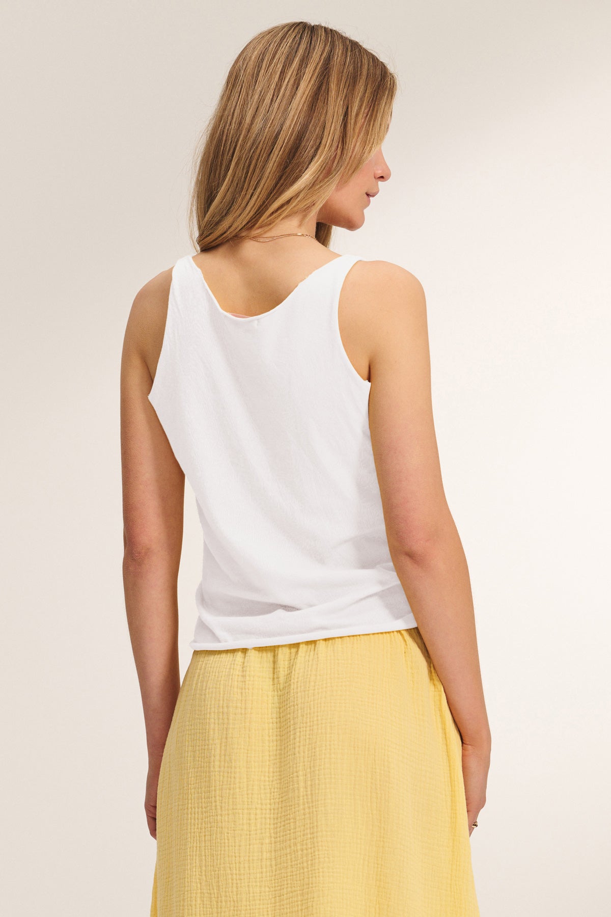 A woman stands with her back to the camera, wearing the Velvet by Graham & Spencer KIM TANK TOP, a white cotton tank with a scoop neckline, paired with a yellow skirt—a true wardrobe essential.-40014168981697