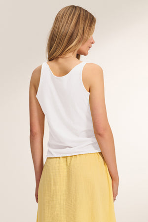 A woman stands with her back to the camera, wearing the Velvet by Graham & Spencer KIM TANK TOP, a white cotton tank with a scoop neckline, paired with a yellow skirt—a true wardrobe essential.