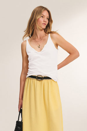 A woman wears the KIM TANK TOP by Velvet by Graham & Spencer—a cotton scoop-neck essential—paired with a yellow skirt, black belt, and woven black bag, posing against a neutral background.