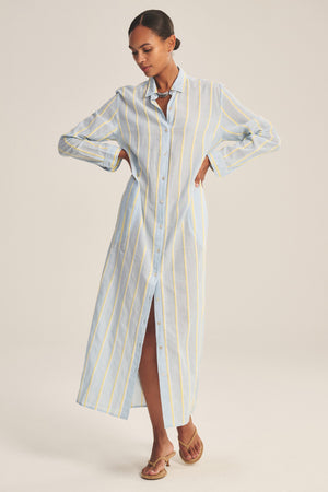 Woman wearing the KIMORA DRESS by Velvet by Graham & Spencer—a long, button-down light blue cotton dress with yellow and white vertical stripes, standing with arms slightly bent and looking down.
