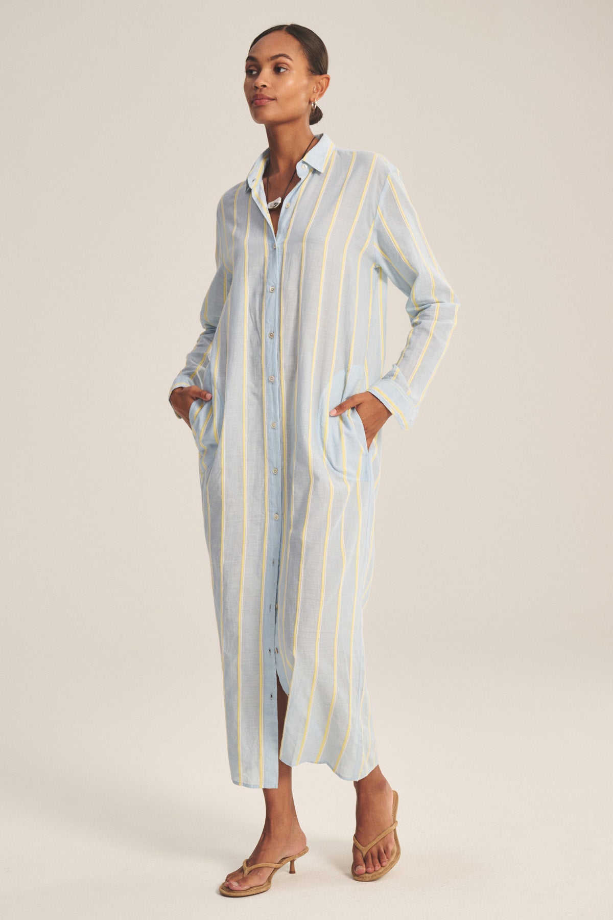 A woman wears the KIMORA DRESS by Velvet by Graham & Spencer, a long cotton dress with vertical yellow and white stripes, button front, pockets, and brown heeled sandals.-41008628105409