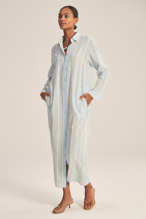 A woman wears the KIMORA DRESS by Velvet by Graham & Spencer, a long cotton dress with vertical yellow and white stripes, button front, pockets, and brown heeled sandals.