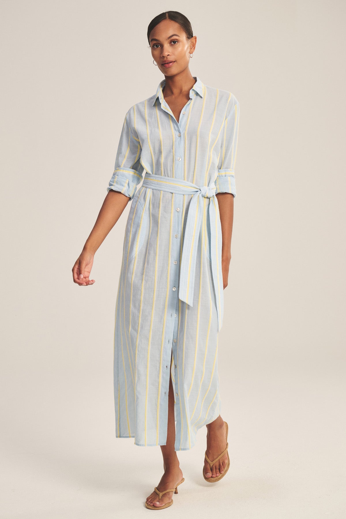 A woman models the KIMORA DRESS by Velvet by Graham & Spencer—a long, button-down cotton stripe dress with a detachable belt and rolled-up sleeves—styled with brown sandals against a plain background.-41008628170945