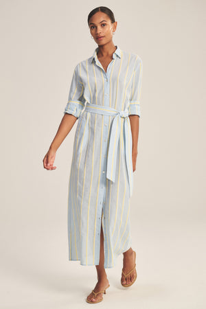A woman models the KIMORA DRESS by Velvet by Graham & Spencer—a long, button-down cotton stripe dress with a detachable belt and rolled-up sleeves—styled with brown sandals against a plain background.