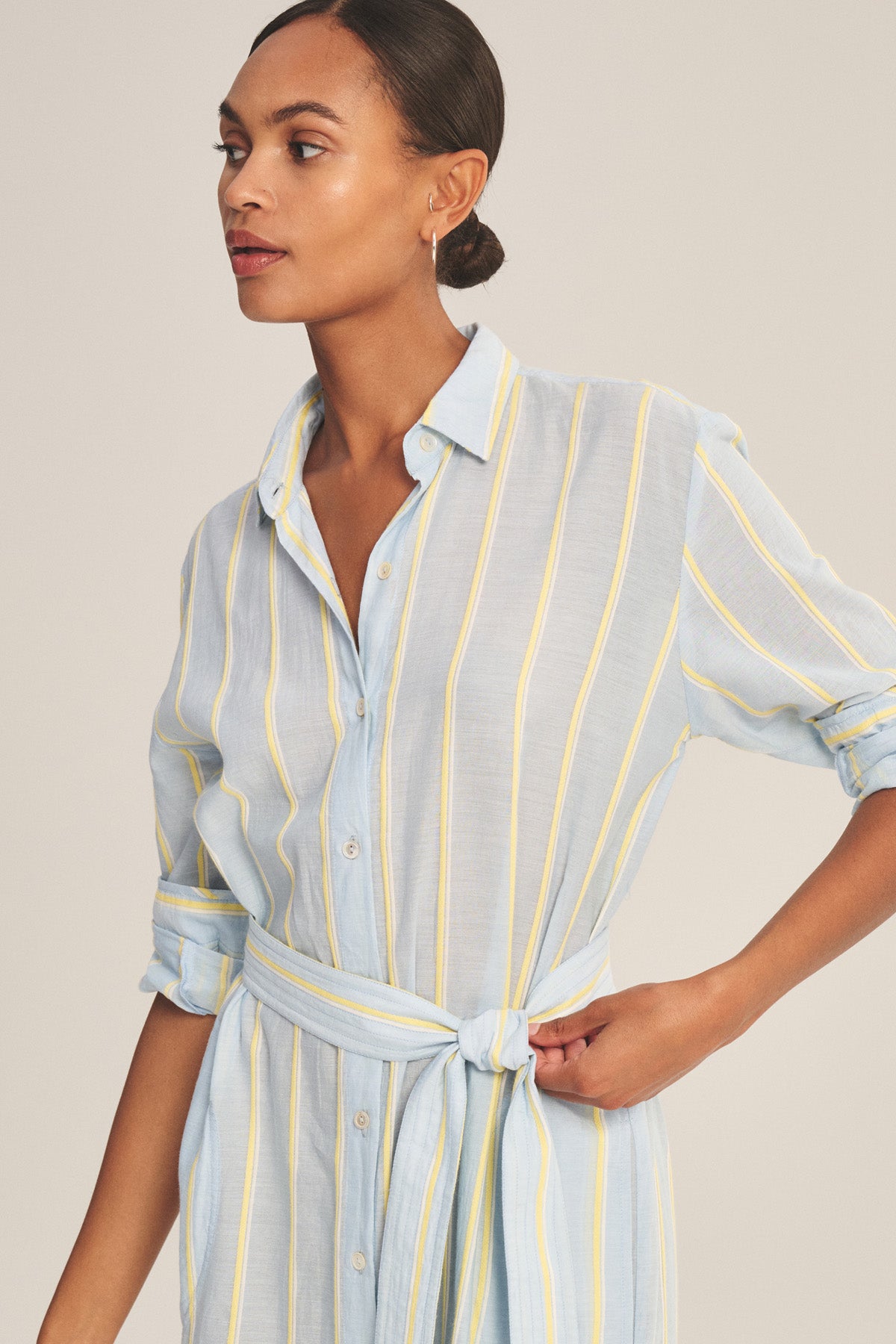 A woman stands with one hand on her hip, looking to the side, wearing the KIMORA DRESS by Velvet by Graham & Spencer—a light blue, yellow, and white striped cotton dress featuring a button front and waist tie.