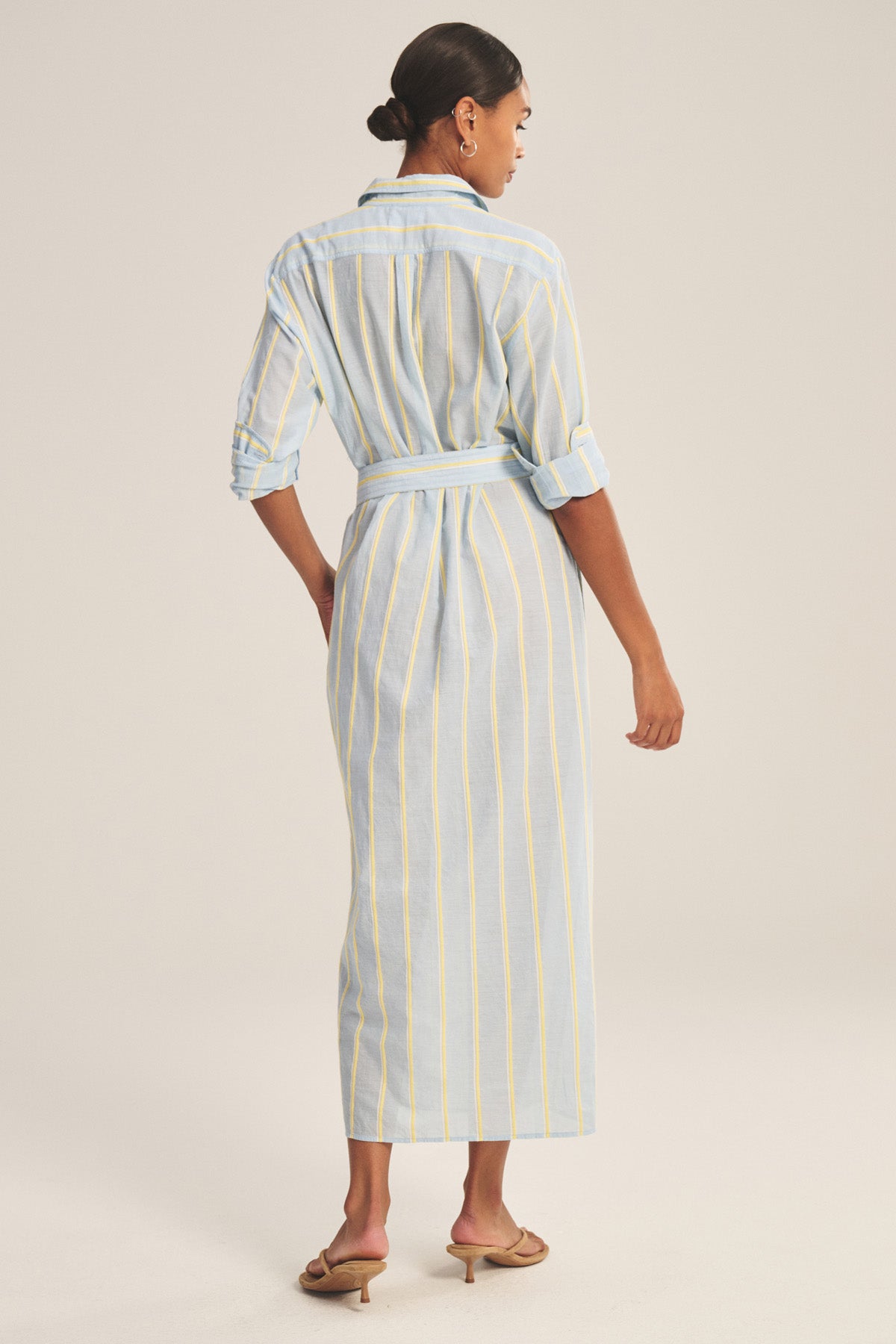 A woman stands facing away, wearing the KIMORA DRESS by Velvet by Graham & Spencer— a light blue cotton maxi dress with yellow and white stripes, detachable belt, rolled-up sleeves, and sandals.