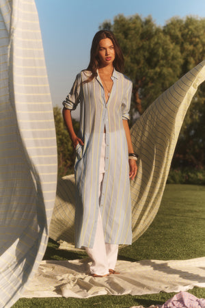 A woman stands outdoors on a striped cloth, wearing the KIMORA DRESS by Velvet by Graham & Spencer over white pants, with fabric panels around her and trees in the background.