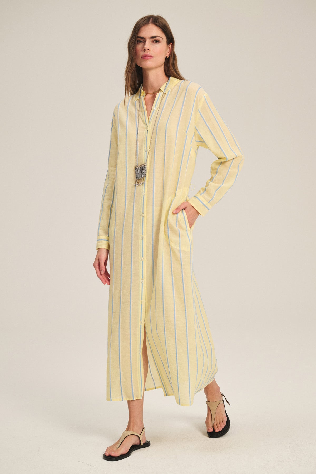 A woman wears the KIMORA DRESS by Velvet by Graham & Spencer—a yarn-dyed cotton dress in light yellow with blue stripes, side pockets, and a detachable belt—paired with flat sandals and a long necklace, against a plain background.-41436017590465
