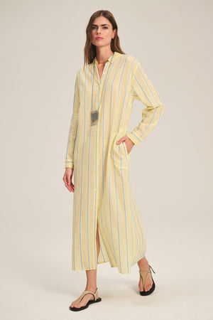 A woman wears the KIMORA DRESS by Velvet by Graham & Spencer—a yarn-dyed cotton dress in light yellow with blue stripes, side pockets, and a detachable belt—paired with flat sandals and a long necklace, against a plain background.