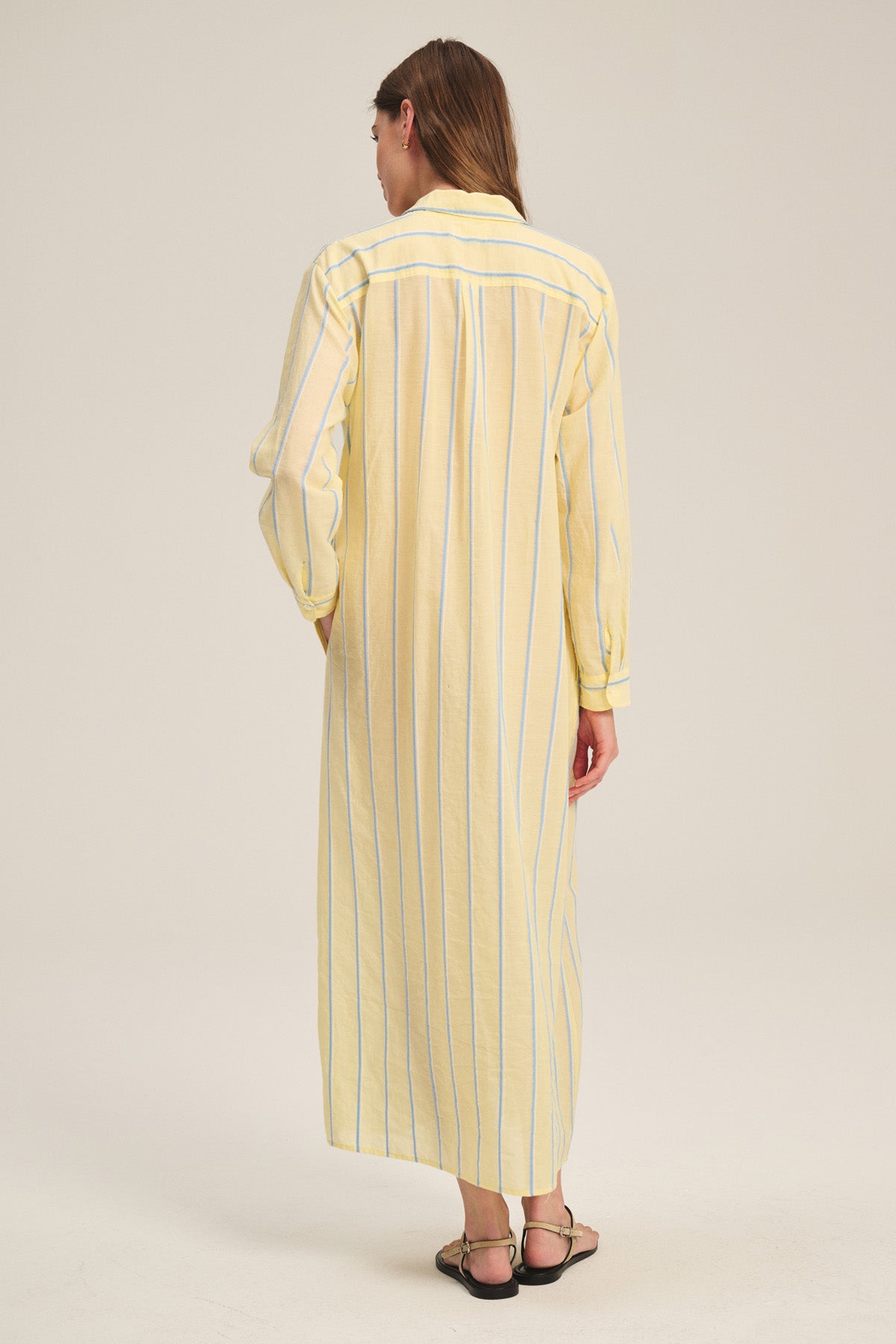 A person faces away, wearing the KIMORA DRESS by Velvet by Graham & Spencer—a long, yellow and white vertically striped cotton maxi dress with long sleeves—paired with black sandals.