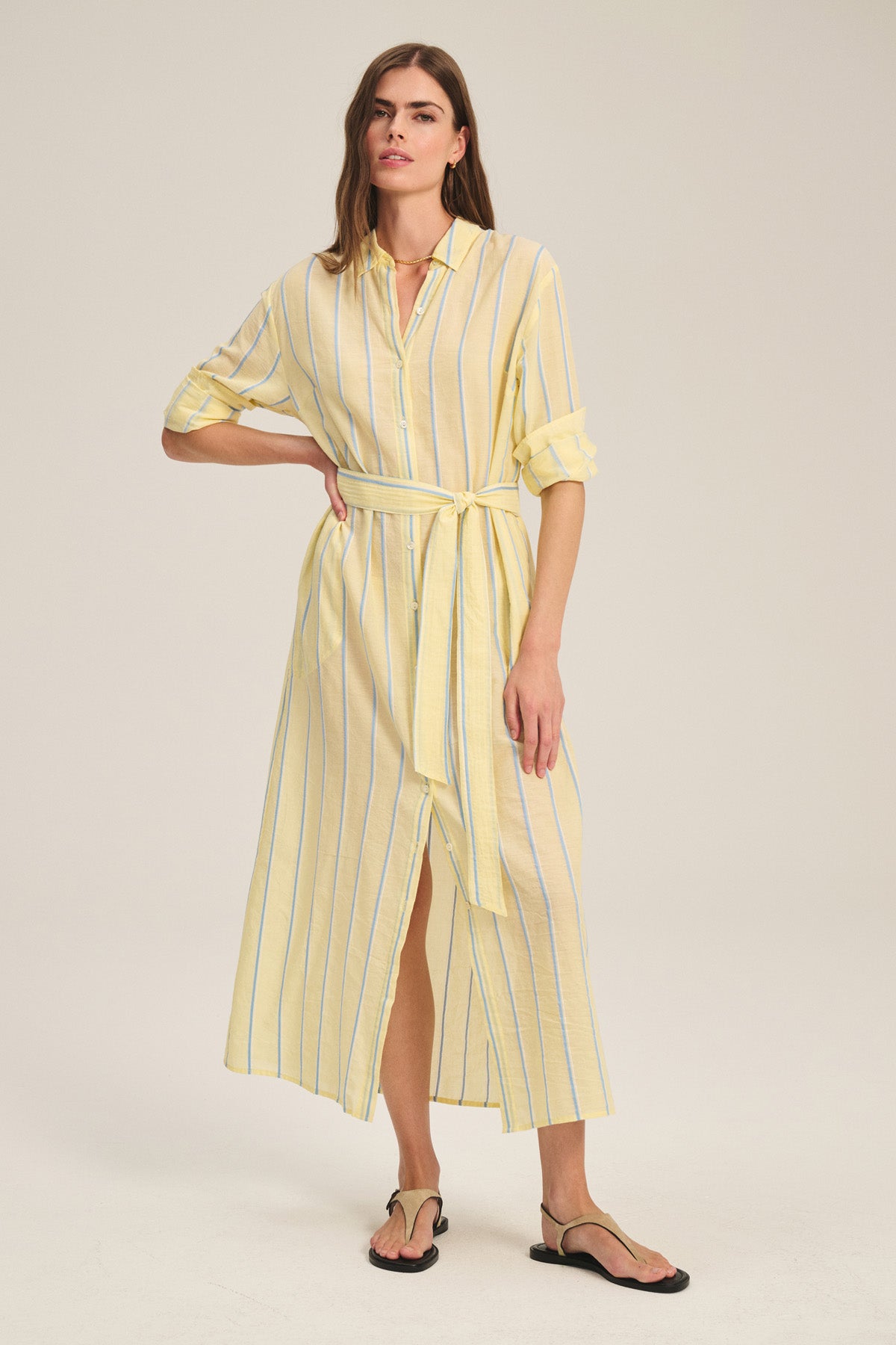 A woman wears the Velvet by Graham & Spencer KIMORA DRESS—a maxi-length, light yellow yarn-dyed stripe cotton dress with rolled-up sleeves and a detachable belt—paired with black sandals against a plain background.-41436017623233