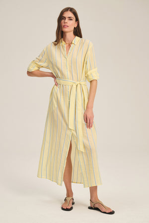 A woman wears the Velvet by Graham & Spencer KIMORA DRESS—a maxi-length, light yellow yarn-dyed stripe cotton dress with rolled-up sleeves and a detachable belt—paired with black sandals against a plain background.