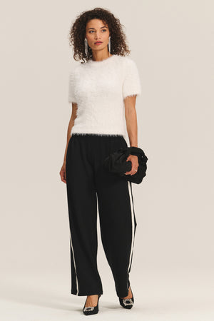 A woman stands against a plain background, wearing the KIT SWEATER by Velvet by Graham & Spencer in white textured fabric, paired with black wide-leg pants with white side stripes, black heels, and holding a black clutch.