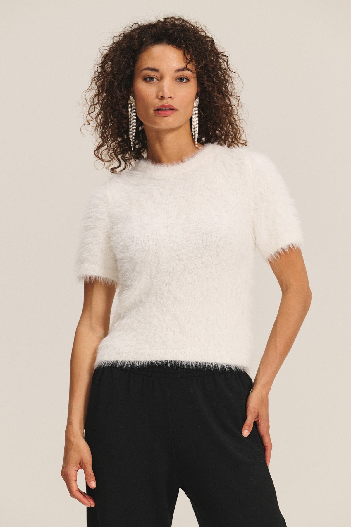 A woman with curly hair wears the KIT SWEATER by Velvet by Graham & Spencer, featuring short sleeves and feather yarn, paired with black pants against a plain background.-40263651557569