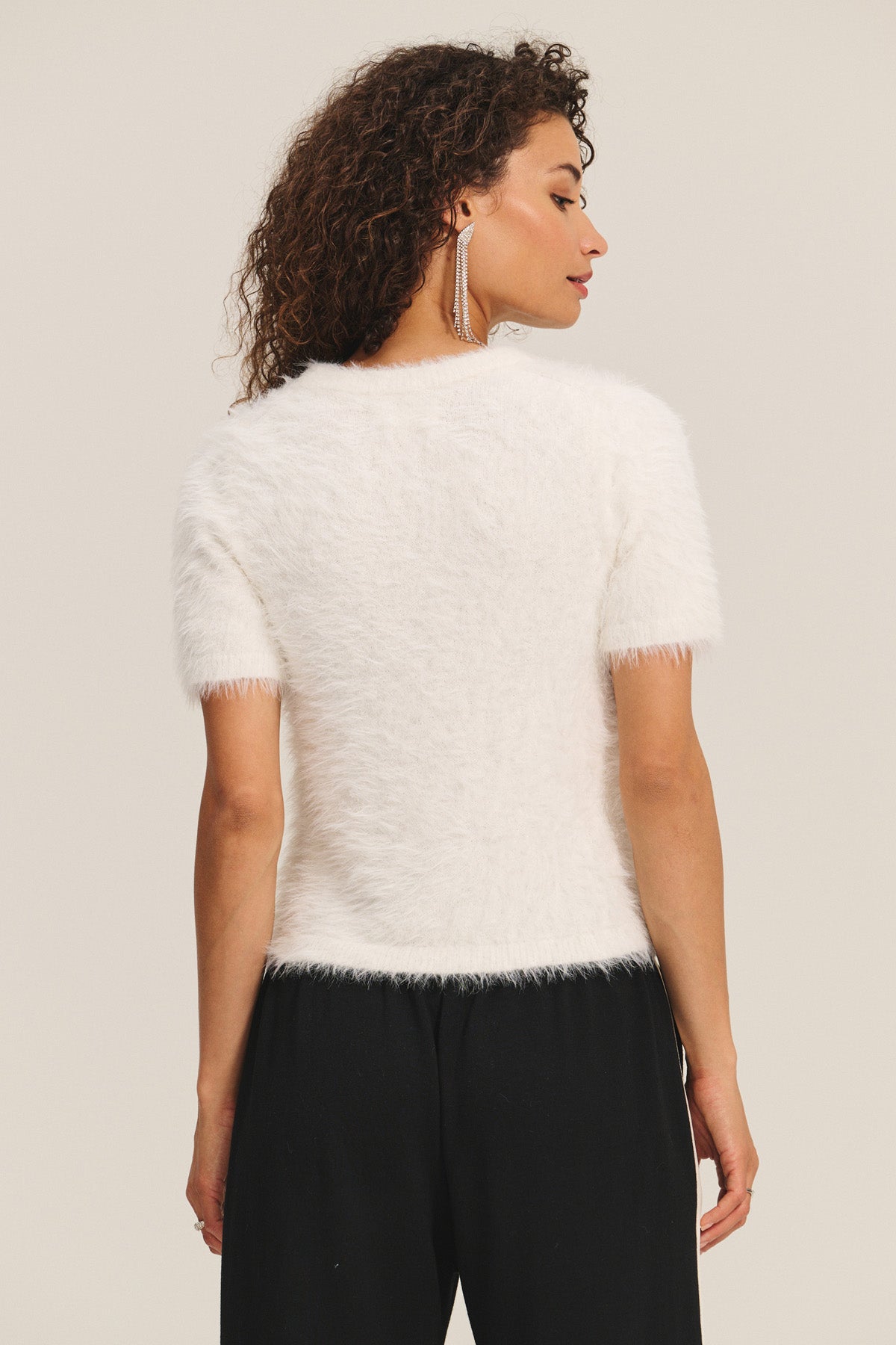 A woman with curly hair is seen from the back wearing the KIT SWEATER by Velvet by Graham & Spencer, a white feather yarn short-sleeve sweater for lightweight comfort, styled with black pants against a plain background.