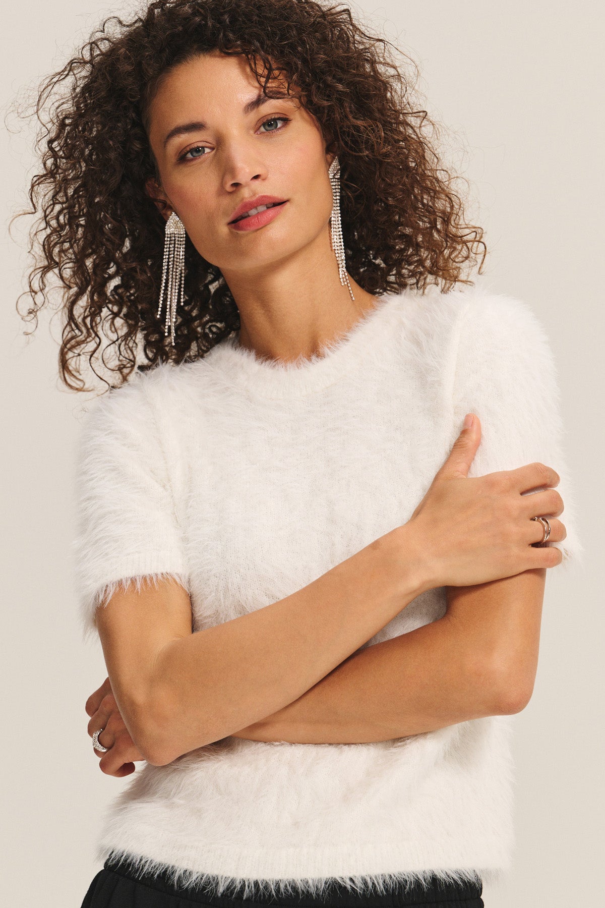 A woman with curly hair crosses her arms in the KIT SWEATER by Velvet by Graham & Spencer—an effortless, lightweight white feather yarn short-sleeve style—paired with dangling earrings and a ring against a plain background.-40263651492033