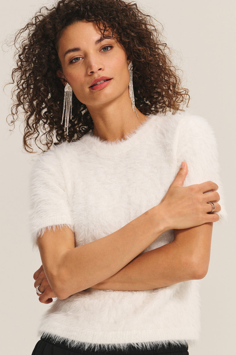 A woman with curly hair crosses her arms in the KIT SWEATER by Velvet by Graham & Spencer—an effortless, lightweight white feather yarn short-sleeve style—paired with dangling earrings and a ring against a plain background.