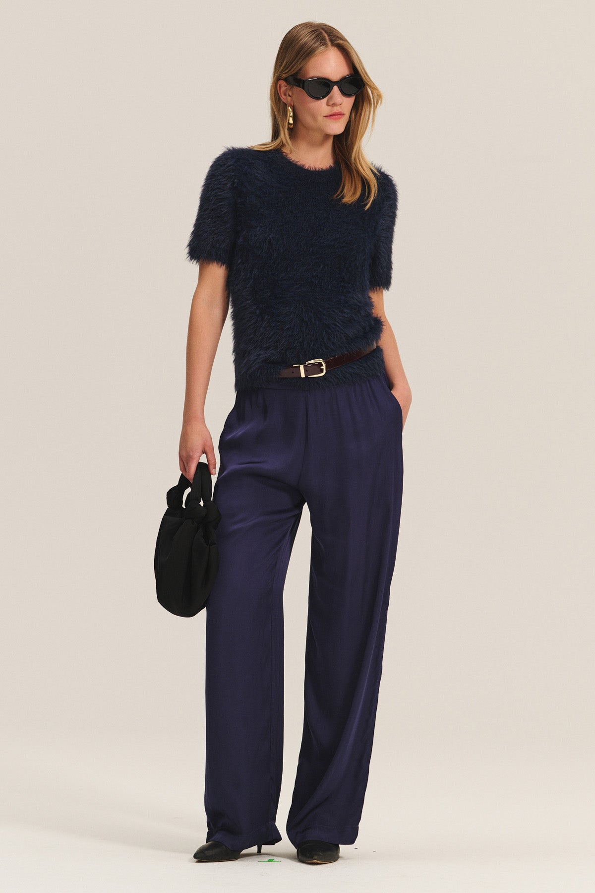 A woman stands against a plain background wearing the KIT SWEATER by Velvet by Graham & Spencer, paired with loose navy blue satin pants, black heeled shoes, sunglasses, and holding a black handbag.-40263651459265