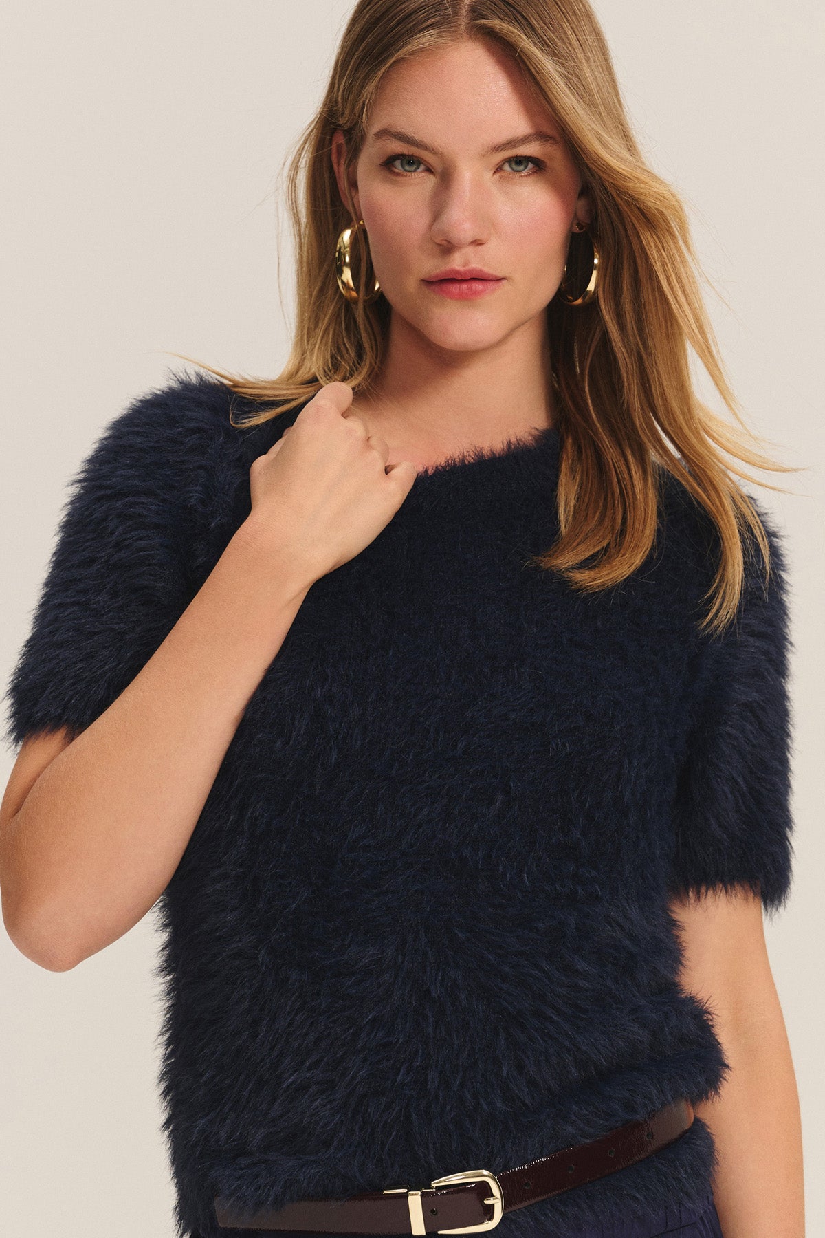 A woman with long blonde hair models the KIT SWEATER by Velvet by Graham & Spencer—a navy feather yarn short-sleeve sweater styled with gold hoop earrings and a dark belt, offering lightweight comfort and effortless style against a plain background.