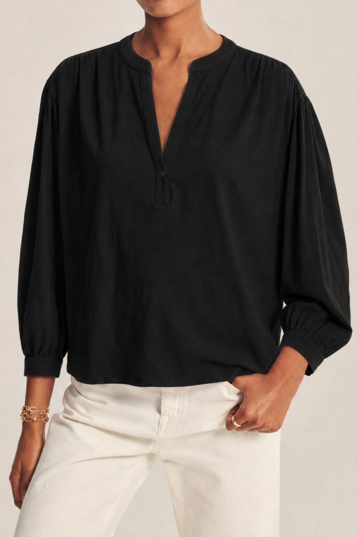 A person is wearing the KNOX TEE by Velvet by Graham & Spencer—a loose-fitting black long-sleeve blouse with a relaxed swing top silhouette and split neckline—paired with cream-colored pants and standing with one hand in a pocket.-42058634428609